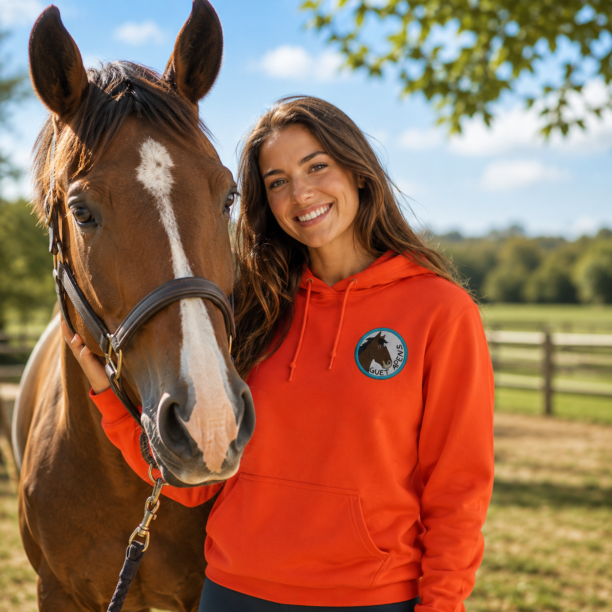 Cavalière souriante portant un sweat à capuche orange personnalisé cheval Guet Apens aux côtés de son cheval dans un paddock en extérieur