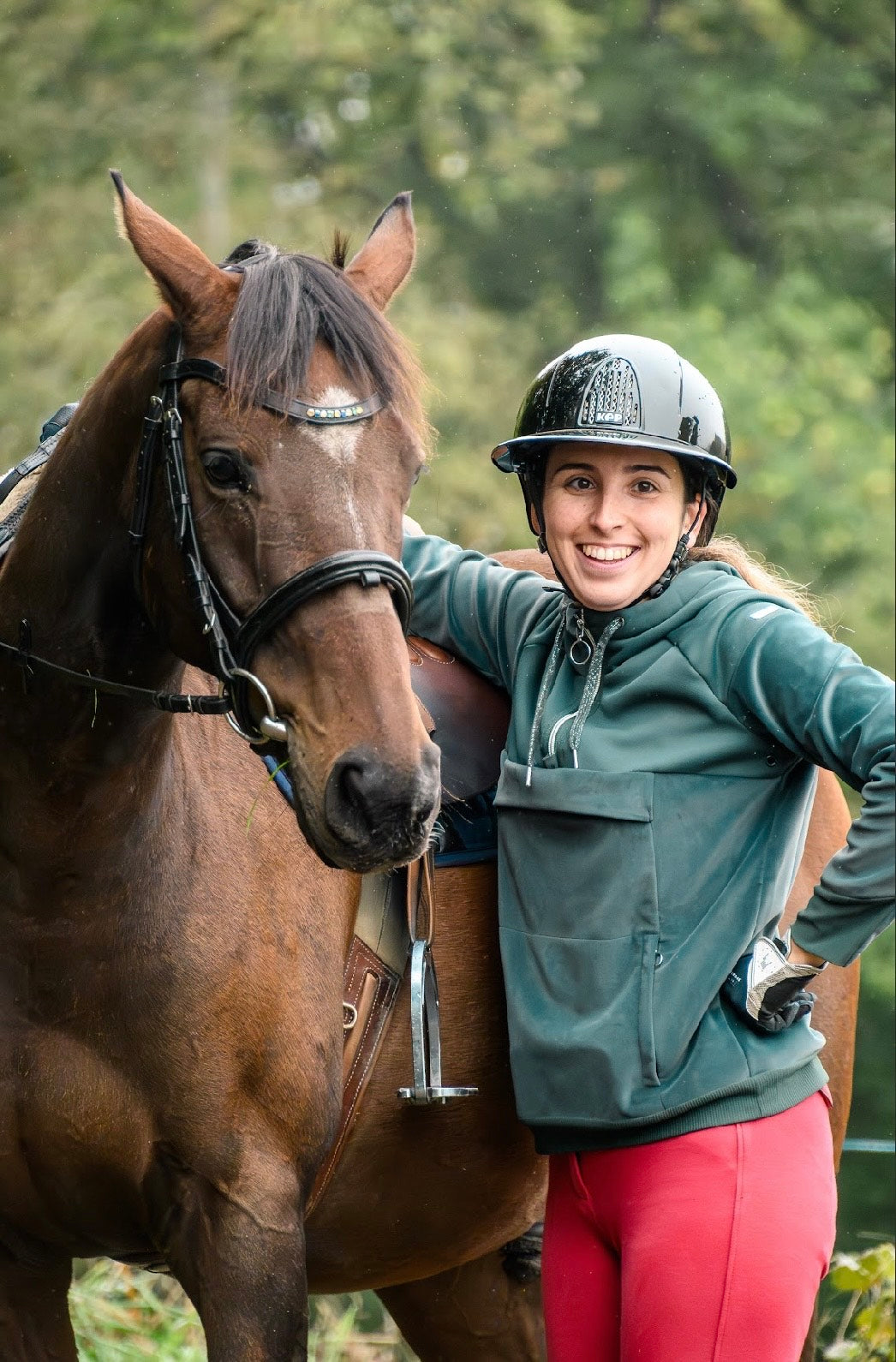 Image représentant Gryffondor des Ombres, un cheval bai, aux côtés de sa cavalière Mathilde, jeune femme blonde et souriante. Leur relation fusionnelle et leur pratique du dressage illustrent le lien fort entre l’animal et sa gardienne. Mathilde est ambassadrice de la marque Libellule Graphisme.